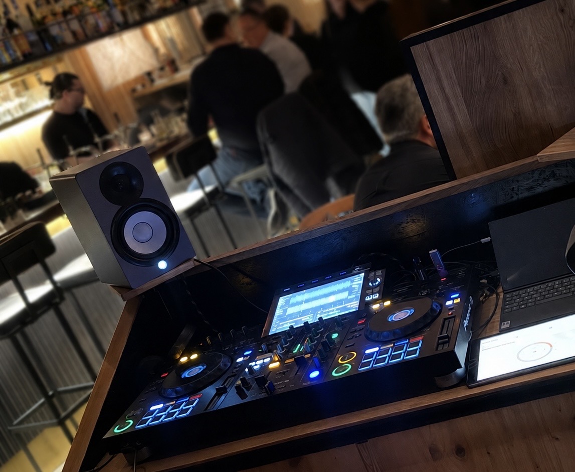 DJ Pult mit blau leuchtenden Reglern in Holzbar mit Gaesten im Hintergrund
